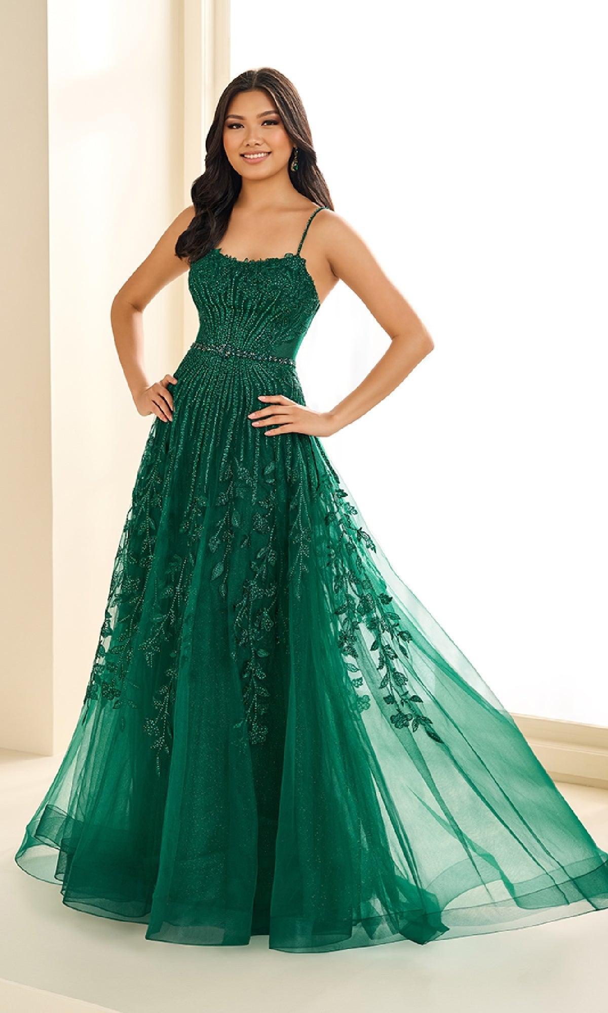 A woman stands indoors wearing the Ellie Wilde EW36068, a floor-length emerald green prom dress with a fitted bodice, thin straps, sleeveless design, and embroidered skirt details.